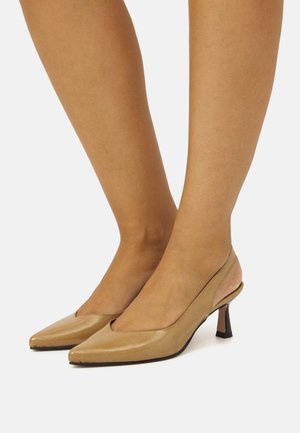 Piedi che indossano décolleté beige con punta affusolata e tacco a blocco curvo basso su uno sfondo bianco.