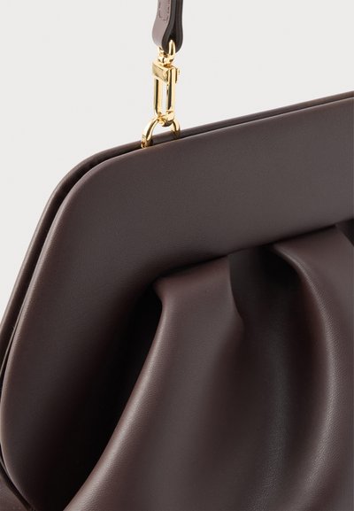 Σκούρο καφέ γωνία δερμάτινης τσάντας με κούμπωμα σε χρυσή απόχρωση και μέρος του προσαρτημένου λουριού σε φωτεινό φόντο.