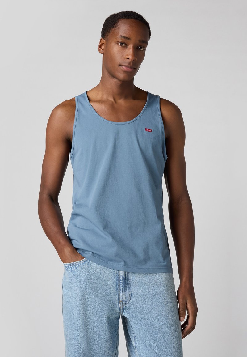 Un débardeur en coton bleu clair avec un col rond et un petit logo rouge sur la poitrine, associé à un jean en denim clair.