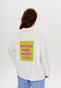 Weißer übergroßer Hoodie mit einem grünen grafischen Rechteck auf dem Rücken, das pinken Text zeigt: "MATCHA MEHR, SORGE WENIGER!"