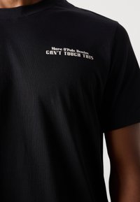 Schwarzes Baumwoll-T-Shirt mit Rundhalsausschnitt und kurzen Ärmeln. Auf der linken Brust steht der Text "Marc O'Polo Denim CAN'T TOUCH THIS" in weiß.