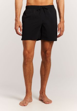 Muški model nosi crne kupaće gaće s elastičnim pojasom i vezicom, stoji bos na jednostavnoj svijetloj pozadini.