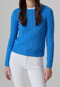 Blauer, kabelgestrickter Pullover mit geripptem Saum und Bündchen. Über einem weißen Hemd getragen, kombiniert mit weißen Hosen. Glatte Textur und taillierte Passform.