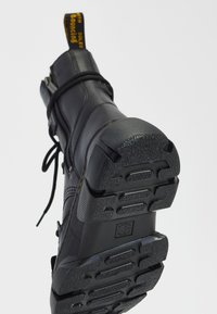 Black leather boot with textured sole, prominent yellow pull tab, and laces. Features stitching details. Bottom shows grip patterns for traction.