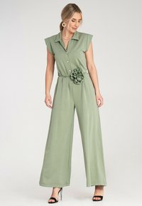 Zielony, bezrękawnikowy kombinezon z kołnierzykiem, zapinany na guziki, o szerokich nogawkach i z akcentem w postaci materiałowego kwiatka w talii. Miękka tekstura i luźny krój.