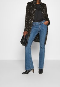 Leopardmönstrad lång kappa, svart blus, blå utsvängda jeans och svarta spetsiga klackar. Hand som håller en svart kuvertväska.