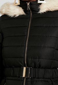 Černá péřová bunda s kapucí s umělým kožíškem, prošitá textura, kovový zip a lesklá stříbrná přezka na opasku.