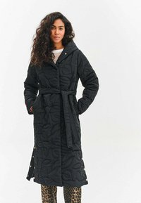 Žena s dlhými kučeravými vlasmi v čiernom prešívanom páse s kapucňou a nohavicami s leopardím vzorom, stojaca na bielom pozadí.