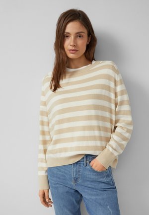 Jonge vrouw met bruin haar, gekleed in een beige-wit gestreepte trui en blauwe spijkerbroek, staand met één hand in de zak tegen een grijze muur.