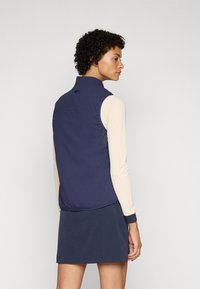 Colete leve azul-marinho com colarinho alto, costas franzidas e mangas longas brancas, usado sobre uma saia azul-marinho correspondente. Design minimalista.