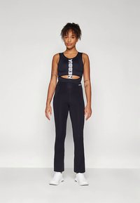Fekete sportmelltartó kivágott dizájnnal és fehér NEBBIA logóval, fekete, testhezálló leggingsszel párosítva. A modell fehér sportcipőt visel.
