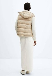 Beige Steppweste mit Kapuze und horizontaler Steppung, getragen über einem weißen strukturierten langen Kleid, kombiniert mit hellen Schuhen.