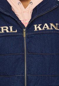 Jeanspuffjacka i marinblått med beige broderad "KARL KANI"-logotyp, gyllene dragkedja, vadderad textur och horisontella sömadderingar.