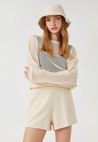 Suéter de malla en crema sobre una camiseta negra, combinado con pantalones cortos de tela color marrón claro. Un sombrero tipo bucket en crema completa el look. Textura suave, ajuste casual.