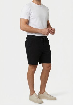 Uomo che indossa una t-shirt bianca a maniche corte, pantaloncini neri con coulisse e sneakers beige, in piedi contro uno sfondo bianco uniforme.