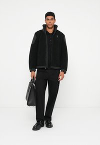 Czarna kurtka polarowa na zamek z przodu, w połączeniu z czarną koszulką polo, czarnymi spodniami i czarną torebką.