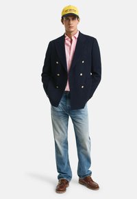 Marine-blauwe gestreepte dubbelgeknoopte blazer, lichtroze shirt, blauwe spijkerbroek en bruin leren schoenen. Gele baseballpet met logo.