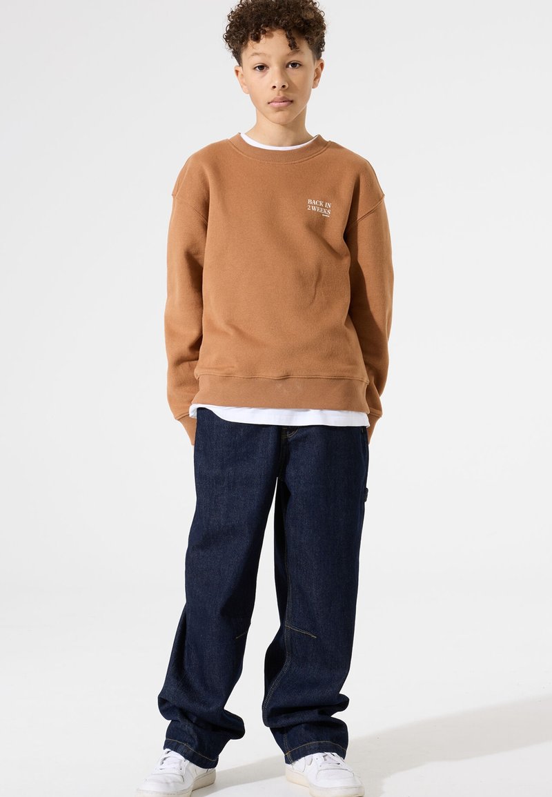 Felpa di cotone marrone con scritta bianca "TORNANDO IN 2 SETTIMANE," abbinata a jeans blu scuro e scarpe da ginnastica bianche.