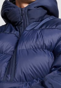 Granatowa kurtka puchowa z wysokim kołnierzem, zamkiem z przodu, pikowanym wzorem i teksturowaną tkaniną. Charakteryzuje się błyszczącym wykończeniem i ociepleniem.