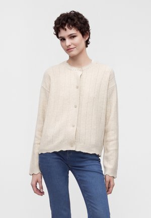 Jeune femme aux cheveux courts et bouclés portant un cardigan crème boutonné et un jean bleu sur un fond blanc uni.