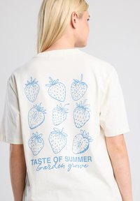 Lichtcrème t-shirt met een blauwe illustratie van aardbeien en de tekst "TASTE OF SUMMER" en "Garden groove" op de achterkant.
