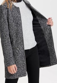 Cappotto grigio e nero con motivo a pattern, dal taglio dritto, zip frontali con tasche e fodera interna nera, indossato aperto sopra un maglione grigio.