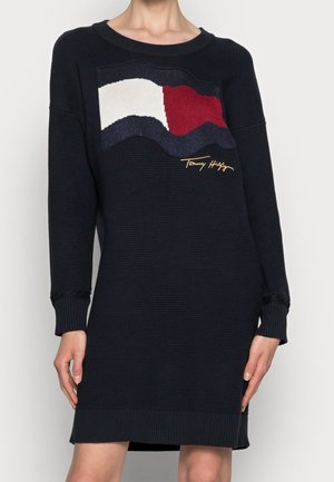 Femme portant une robe en maille noire avec un motif en vagues rouge, blanc et marine et une signature jaune sur la poitrine.