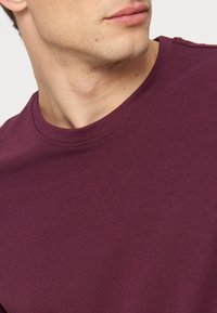 Κοντινό πλάνο ενός άνδρα που φοράει ένα μονόχρωμο σκούρο μοβ t-shirt με στρογγυλή λαιμόκοψη, κοιτάζοντας προς το πλάι με ορατό μόνο το κάτω μέρος του προσώπου και τον λαιμό.