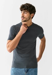 Vīrietis ar cirtainiem matiem un bārdu, ģērbies pieguļošā tumši pelēkā T-kreklā un zilās džinsās, skatās uz sāniem ar vienu roku pie zoda.