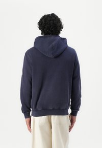 Uomo con capelli neri ricci, indossa una felpa blu navy e pantaloni beige, che guarda di spalle su uno sfondo bianco uniforme.