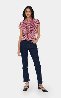 Roza bluzica z naborki s cvetličnim vzorcem, v kombinaciji z temno modrimi kavbojkami. Model nosi črne čevlje s kvadratnimi prsti in gležnjakom.