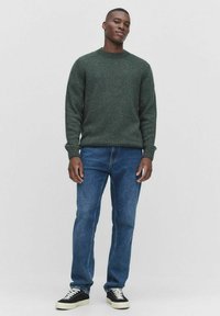 Groene geweven trui met ronde hals, gecombineerd met blauwe rechte jeans en zwarte sneakers met witte accenten.