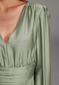 Robe vert olive avec un décolleté en V profond, des manches longues froncées et une taille cintrée avec des plis, fabriquée en tissu lisse.