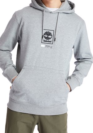 Osoba ubrana w jasnoszarą bluzę Timberland z kapturem, posiadającą przednią kieszeń i ściągacze, w połączeniu z oliwkowymi spodniami.