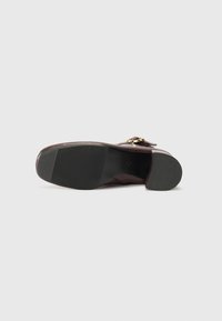 Chaussure en cuir brun au design structuré, dotée d'une semelle texturée, de détails en métal doré, et d'un bout arrondi. Fini supérieur lisse.