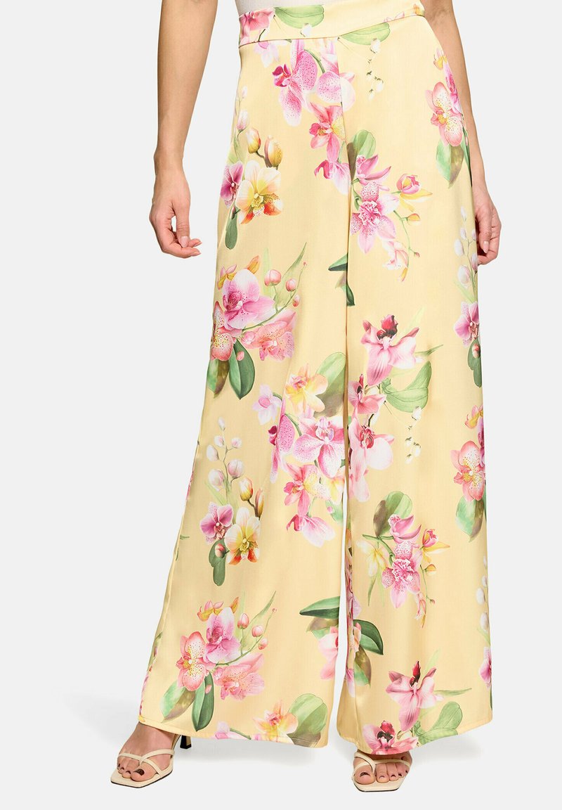 Pantalon jaune à jambes larges avec imprimé floral d’orchidées roses et blanches, porté avec des sandales beiges ouvertes à talons.