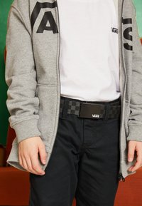 Grå zip-up hoodie med svart logotyp, kombinerad med mörka byxor och ett svart rutigt bälte med en rektangulär spänne.