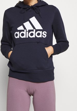 Person trägt einen dunkelblauen Adidas-Hoodie mit weißem Logo und pinken Leggings, die Hände in der vorderen Tasche des Hoodies, steht vor einem einfarbigen Hintergrund.