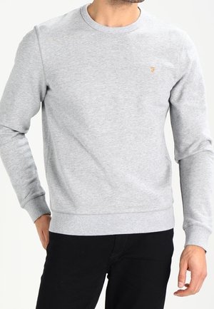 Homme portant un sweat-shirt gris clair à col rond avec manches longues et petit logo brodé sur la poitrine, associé à un pantalon noir.