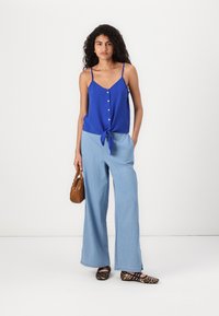 Haut sans manches bleu avec fermeture à boutons à l'avant et détail à nouer, associé à un pantalon large bleu clair et des ballerines à imprimé léopard. Sac à main marron.