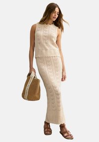Knit two-piece outfit in light beige featuring a sleeveless top and a long skirt with intricate patterns. Accessorized with a woven tote bag.