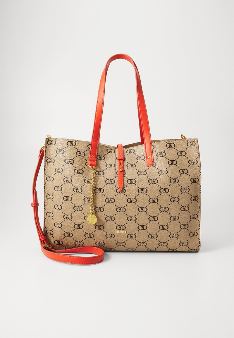 Sac fourre-tout beige avec logo noir à motif, poignées et bandoulière en cuir rouge, et une chaîne dorée avec un pendentif rond suspendu à l'avant.