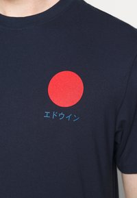 Námořnické bavlněné tričko s velkým červeným kruhem a modrým japonským textem na hrudi. Krátké rukávy a kulatý výstřih.