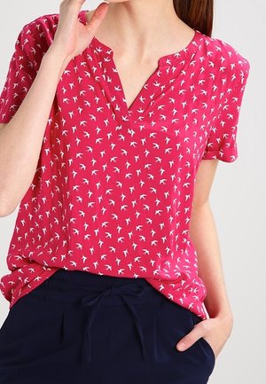 Femme portant un chemisier rose à manches courtes avec des motifs d'oiseaux blancs, rentré dans un pantalon bleu marine à cordon, main reposant dans la poche.