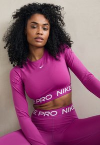 Prilagođena, dugosleevna atletska majica i odgovarajuće visoke tajice u jarkoj ružičastoj boji. Nike logo i "PRO NIKE" oznaka na pojasu. Glatka tekstura.