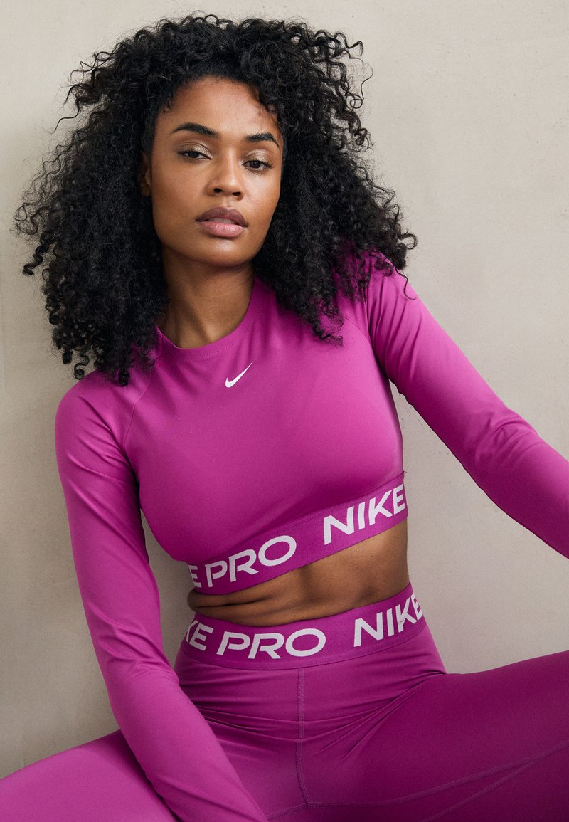 Prilagođena, dugosleevna atletska majica i odgovarajuće visoke tajice u jarkoj ružičastoj boji. Nike logo i "PRO NIKE" oznaka na pojasu. Glatka tekstura.