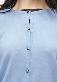 Gros plan sur un cardigan en maille bleu clair avec cinq boutons argentés et brillants attachés le long du devant, porté par une personne aux cheveux foncés.