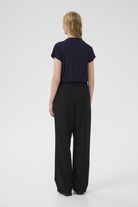 Blondýnka stojící zády ke kameře, oblečená v tmavě modrém krátkém tričku a volných černých kalhotách na bílém pozadí.