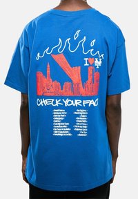 Camiseta azul de algodón con diseño gráfico en la parte posterior: horizonte de ciudad estilizado, llamas y texto que dice “I ❤️ NY” y “REVISA TU ROSTRO.”