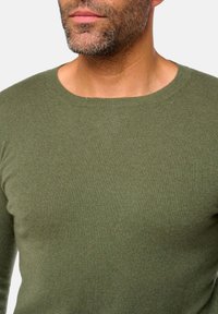Homme avec une barbe courte portant un pull à col rond vert olive devant un fond clair uni.